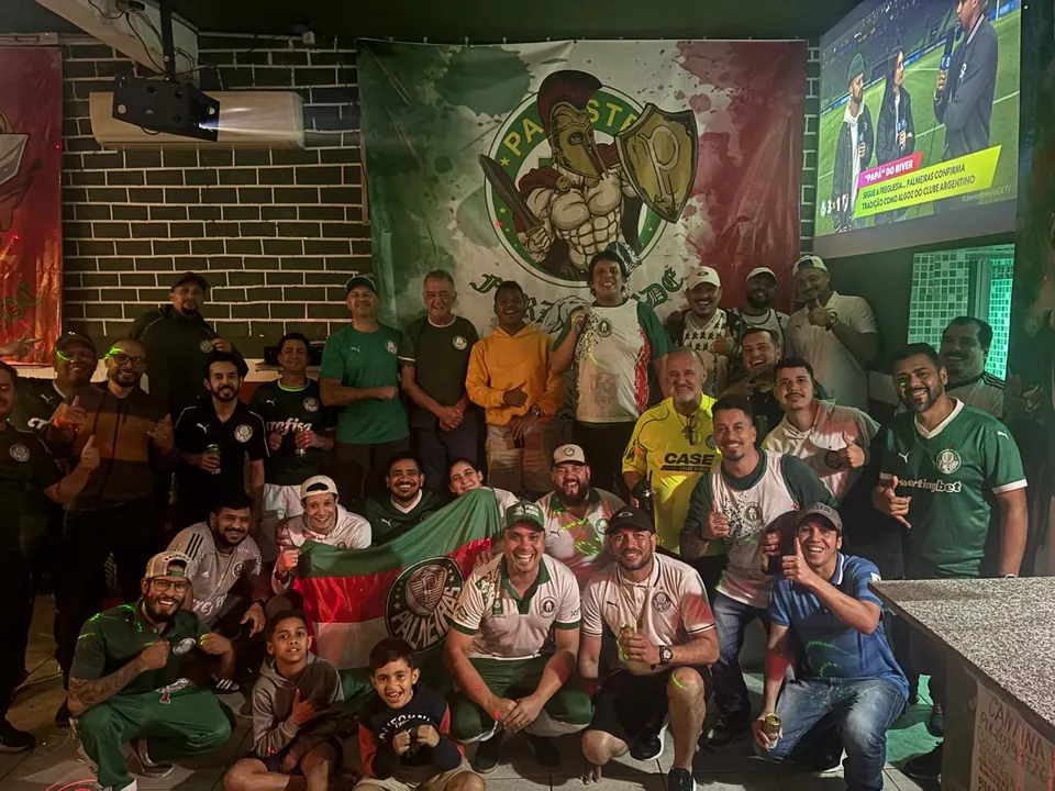 Cerca de trinta torcedores posam no salão da sede, à frente do bandeirão com o espartano da Forza, com o telão ligado à direita e uma bandeira do Palmeiras aberta na frente.