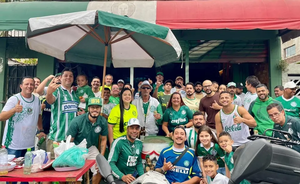 Grupo grande de torcedores, adultos e crianças, com camisas do Palmeiras na calçada em frente à sede, sob o toldo verde, branco e vermelho e um guarda-sol.