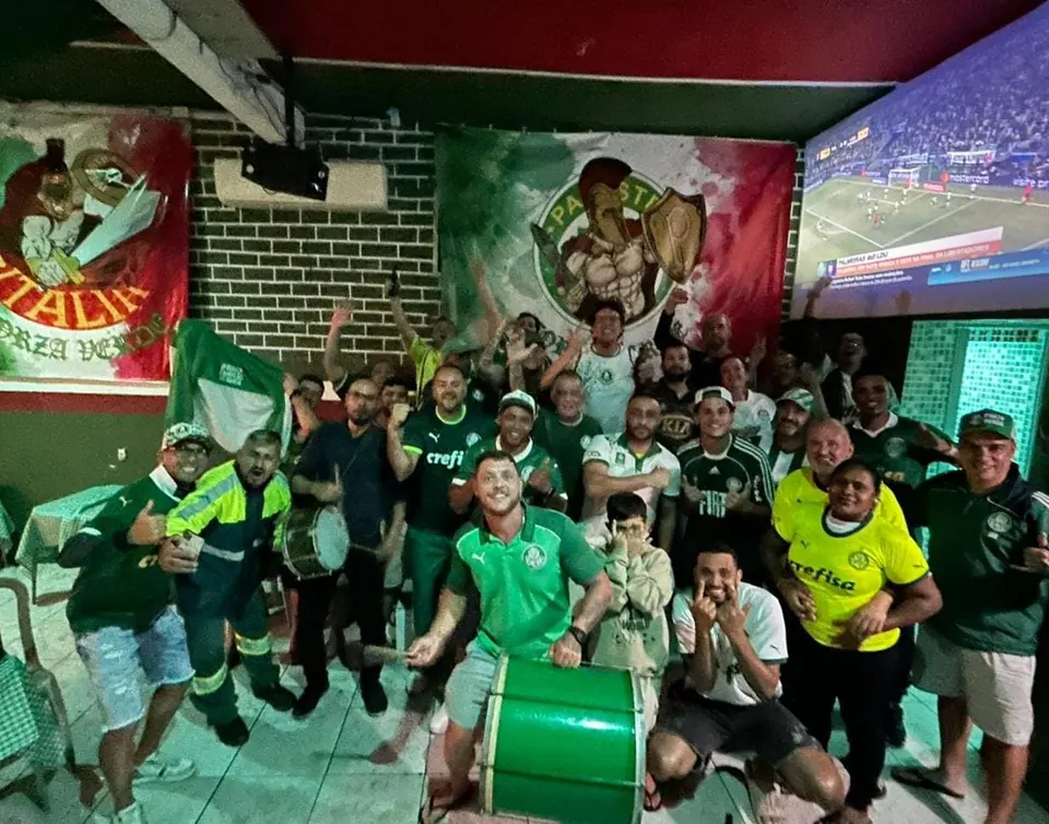 Torcedores comemoram no salão da sede com os braços para cima, um bumbo verde em primeiro plano, o bandeirão do espartano na parede e o jogo passando no telão.